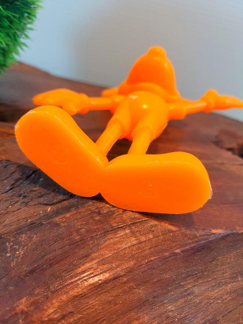 May include: Orange plastic figurine of a cartoon character with large feet and outstretched arms.