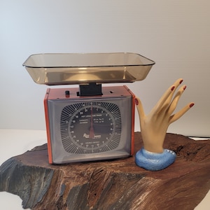 May include: A vintage red and gray kitchen scale with a clear amber-colored tray. The scale has a black face with white numbers and a needle indicator. A decorative hand statue with red nail polish is next to the scale.