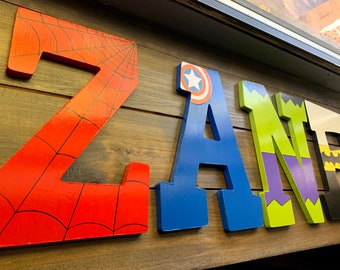May include: Four large wooden letters spelling the name ZANE. The Z is red with a black spiderweb pattern. The A is blue with a circular shield emblem at the top. The N is lime green and purple with jagged edges. The E is black with yellow accents.