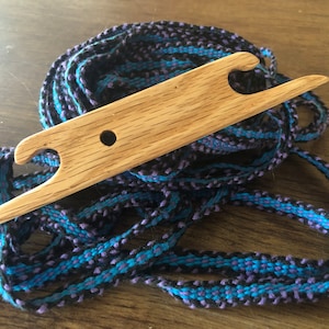 May include: A wooden weaving tool with two holes and a pointed end, resting on a pile of blue and black braided cord.