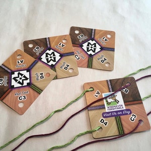 May include: Four square wooden loom cards with holes for string. The cards are labeled with different wood types, including walnut, cherry, red oak, and maple. The cards also have numbers on them, such as A1, B2, C3, and D4. The cards are on a white surface with three strands of string.