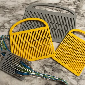 May include: Three rigid heddle weaving tools with arched handles, including one large gray tool, one medium yellow tool, and one small yellow tool. A small gray weaving tool is threaded with blue, green, white, and brown yarn in a woven pattern.
