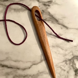 May include: A wooden needle threader with a dark red cord threaded through the eye. The threader is on a white marble surface.