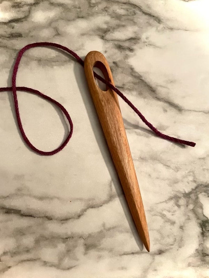 May include: A wooden needle threader with a dark red cord threaded through the eye. The threader is on a white marble surface.