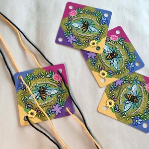 May include: Square tags with a bee and floral design, featuring the text "When the flower blossoms, the bee will come." The tags have a gradient of purple, yellow, and blue. Black and yellow strings are attached.