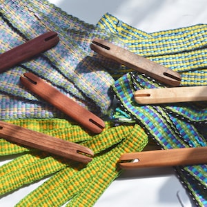 May include: A collection of wooden weaving tools, called heddles, with colorful woven bands. The heddles are made of different types of wood and have a simple design with two holes at each end.