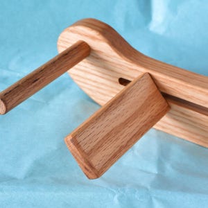 May include: A wooden guitar stand with a natural finish. The stand has a curved base and a vertical support with a slot for the guitar neck. The stand is designed to hold a guitar securely in place.