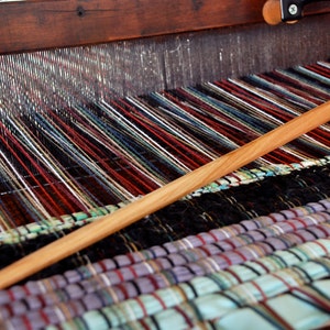 May include: A close-up of a wooden loom with colorful threads woven into a striped pattern. A wooden shuttle is being used to weave the threads.