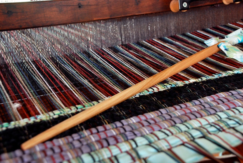 May include: A close-up of a wooden loom with colorful threads woven into a striped pattern. A wooden shuttle is being used to weave the threads.