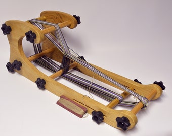 May include: A wooden weaving loom with black knobs and multi-colored yarn. The loom is made of light-colored wood and has a rectangular frame. The yarn is a mix of black, white, green, and purple. A small spool of yarn is also visible.