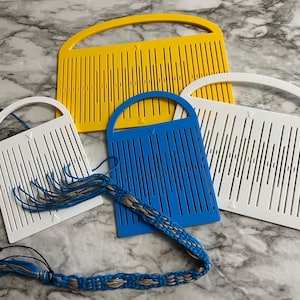 May include: A collection of four rigid heddle weaving tools in yellow, blue, and white. These flat, rectangular plastic tools feature vertical slots and holes for threading yarn, with arched handles at the top and small triangular alignment markers.