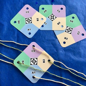 May include: Four diamond-shaped cards with different colored squares and the letters A1, B2, C3, and D4 printed on them. The cards are on a blue surface with white and green yarn.