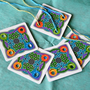 May include: A collection of square game cards with a Celtic knot design in green, purple, and blue, each with colorful rings. Strands of blue and yellow thread are attached to the cards, suggesting a craft or game. The cards are arranged on a teal surface.
