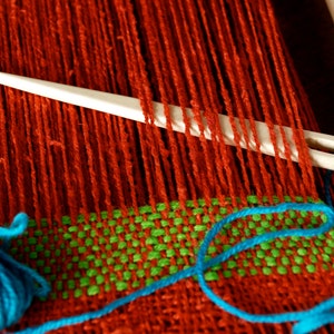 May include: A close-up of a loom with red, green, and blue yarn. A white plastic needle is threaded through the red yarn.