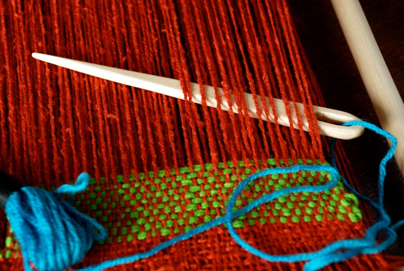 May include: A close-up of a loom with red, green, and blue yarn. A white plastic needle is threaded through the red yarn.