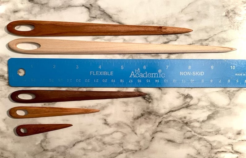 May include: Four wooden yarn needles of varying lengths, with a blue ruler measuring up to 10 inches. The needles are all brown and have a small loop at one end.