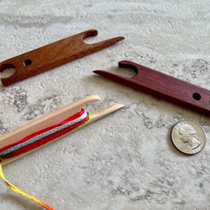 May include: Three wooden yarn winders, two are dark brown and one is light wood. The light wood winder has a multi-colored yarn wound around it. A US quarter is included for scale.