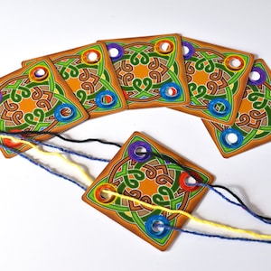 May include: A set of wooden game pieces with a Celtic knot design. Each piece has a central orange circle, surrounded by green and brown knotwork, and colored holes. Strands of yarn are threaded through the holes.