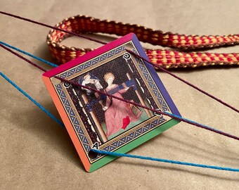 May include: A diamond-shaped card with a medieval illustration, framed in a rainbow border, is strung with blue and purple threads. A woven red and gold band lies in the background. The card features a scene of figures in historical attire.