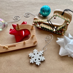 May include: A set of four wooden Christmas ornament holders, each with a different design. The holders are made of natural wood and have a rustic look. The set includes a snowflake, a bow, a blue bauble, and a red bow.