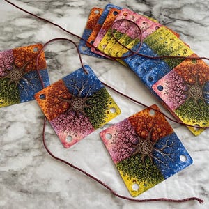 May include: A set of square, multi-colored cards featuring a four-season tree design with a central Celtic knot. Each card has four corner holes and is strung together with a thin, dark red cord, displaying vibrant orange, pink, green, and blue hues.