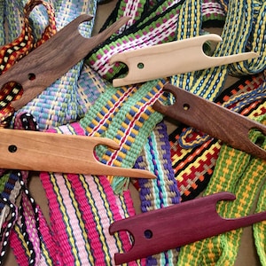May include: Five wooden weaving tools, called pick-up sticks, are arranged on a brown surface. The tools are made of different types of wood and have a variety of colors, including brown, light brown, and purple. The tools are surrounded by colorful woven bands.