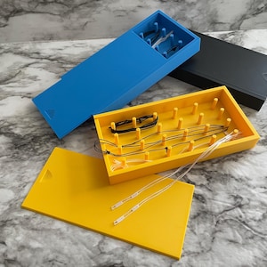 May include: Three rectangular storage boxes in blue, black, and yellow, each featuring a sliding lid. The yellow box is open, revealing a grid of vertical pegs used for organizing thin black and clear cords or strings for jewelry or crafts.