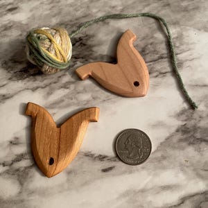 May include: Two wooden yarn holders shaped like fish with a hole in the middle. A ball of green, yellow, and white yarn is next to the holders.