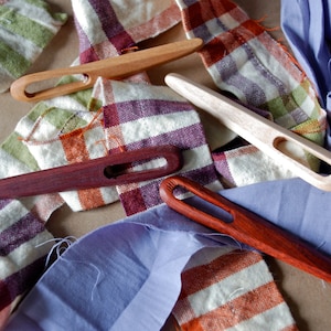 May include: Four wooden and wooden-handled knitting needles with different colored tips, lying on a fabric background. The fabric is a mix of white, orange, purple, and blue.