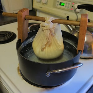 Puede incluir: Una bolsa de tela blanca llena de queso está suspendida sobre una olla de agua hirviendo en una estufa. La bolsa se mantiene en su lugar por un marco de madera con una clavija de madera.