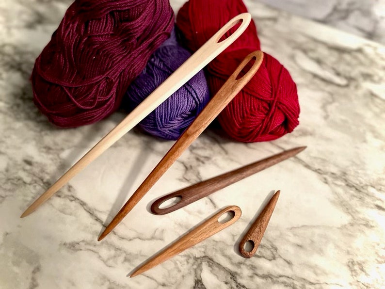 May include: Four wooden yarn needles of varying sizes and colors. The needles are made of light and dark wood and have a small eye at one end. The needles are lying on a white marble surface.