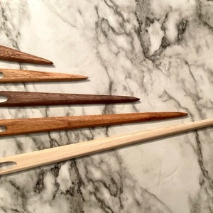 May include: Five wooden knitting needles of varying lengths and colors, arranged in a row on a white marble surface.