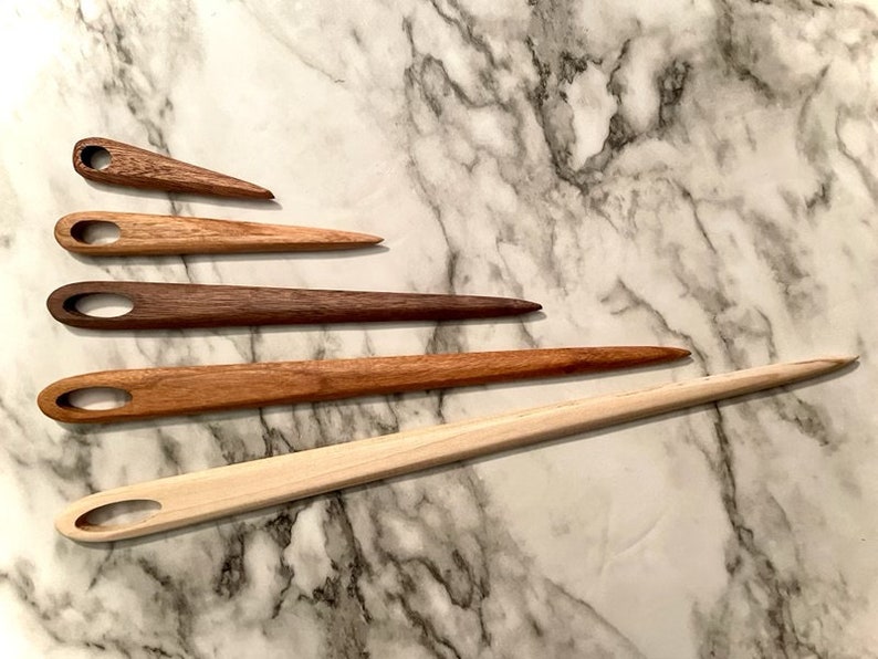 May include: Five wooden knitting needles of varying lengths and colors, arranged in a row on a white marble surface.