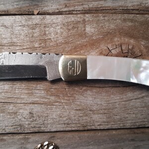 May include: A pocket knife with a white mother-of-pearl handle and a gold monogrammed plate. The blade is made of Damascus steel and has a serrated edge.