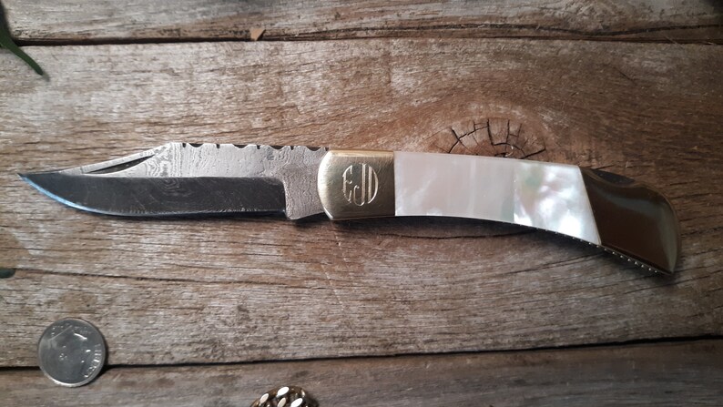 May include: A pocket knife with a white mother-of-pearl handle and a gold monogrammed plate. The blade is made of Damascus steel and has a serrated edge.