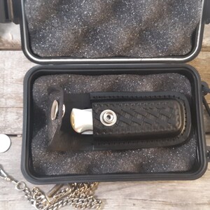May include: A black folding knife with a silver blade is stored in a black hard case with a black strap. The case is open and the knife is visible inside.