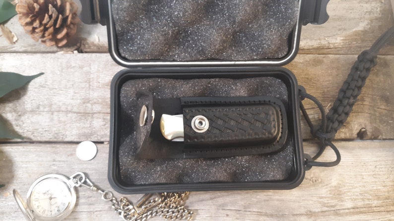 May include: A black folding knife with a silver blade is stored in a black hard case with a black strap. The case is open and the knife is visible inside.