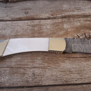 May include: A white and gold pocket knife with a damascus steel blade.