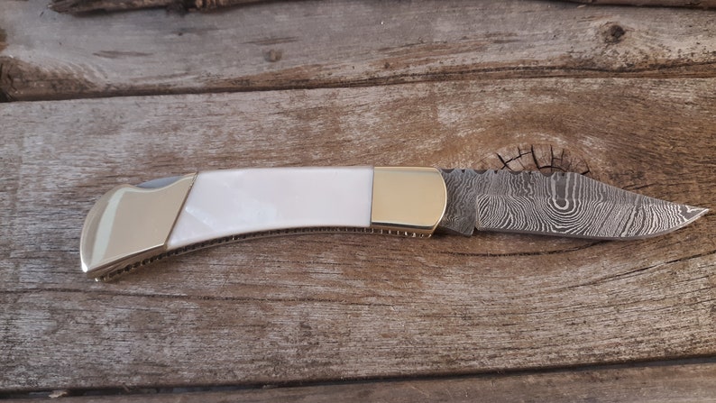 May include: A white and gold pocket knife with a damascus steel blade.