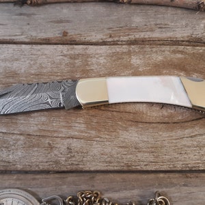 May include: A pocket knife with a white handle and a patterned steel blade. The blade is closed and the knife is lying on a wooden surface.