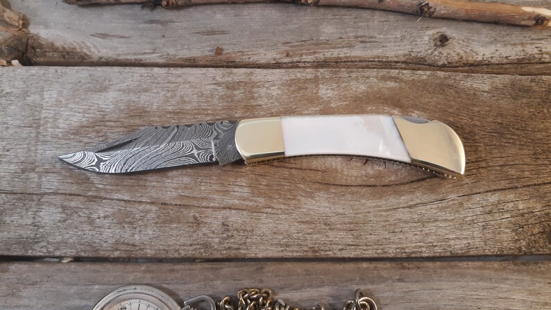 May include: A pocket knife with a white handle and a patterned steel blade. The blade is closed and the knife is lying on a wooden surface.