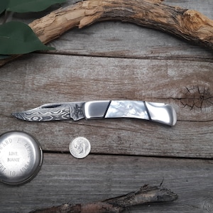 May include: A silver pocket knife with a patterned blade and a white handle. The knife is lying on a wooden surface.
