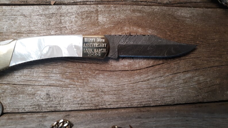 May include: A pocket knife with a white handle and a black blade. The knife has a gold inscription that reads "HAPPY 30th ANNIVERSARY LOVE MARCIE 10-12-93".