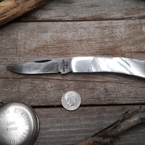 Puede incluir: Un cuchillo de bolsillo plateado con mango de nácar blanco. La hoja está parcialmente abierta y el cuchillo está sobre una superficie de madera.