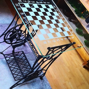 Può includere: Un tavolo da scacchi unico con un piano in vetro con un motivo a scacchiera in bianco e nero. Il tavolo è sostenuto da una base di macchina da cucire nera vintage, che aggiunge un tocco antico. Il tavolo misura circa 76 cm di lunghezza.