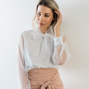 Puede incluir: Una blusa blanca con una superposici&oacute;n de lunares transparentes y una corbata de lazo. La blusa se combina con una falda rosa melocot&oacute;n con un lazo en la cintura.
