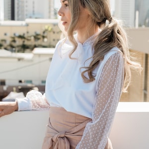 Puede incluir: Una blusa blanca con lazo y mangas transparentes de lunares, combinada con una falda marr&oacute;n claro de cintura alta.