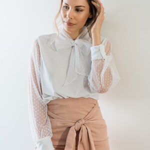 Puede incluir: Una blusa blanca con una superposici&oacute;n de tul de lunares y una corbata de lazo. La blusa se combina con una falda rosa melocot&oacute;n con un lazo en la cintura.