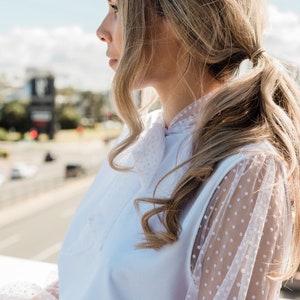 Puede incluir: Una mujer lleva una blusa blanca con una superposici&oacute;n de malla de lunares blancos en las mangas. La blusa tiene un lazo en el cuello. Lleva una falda marr&oacute;n claro.