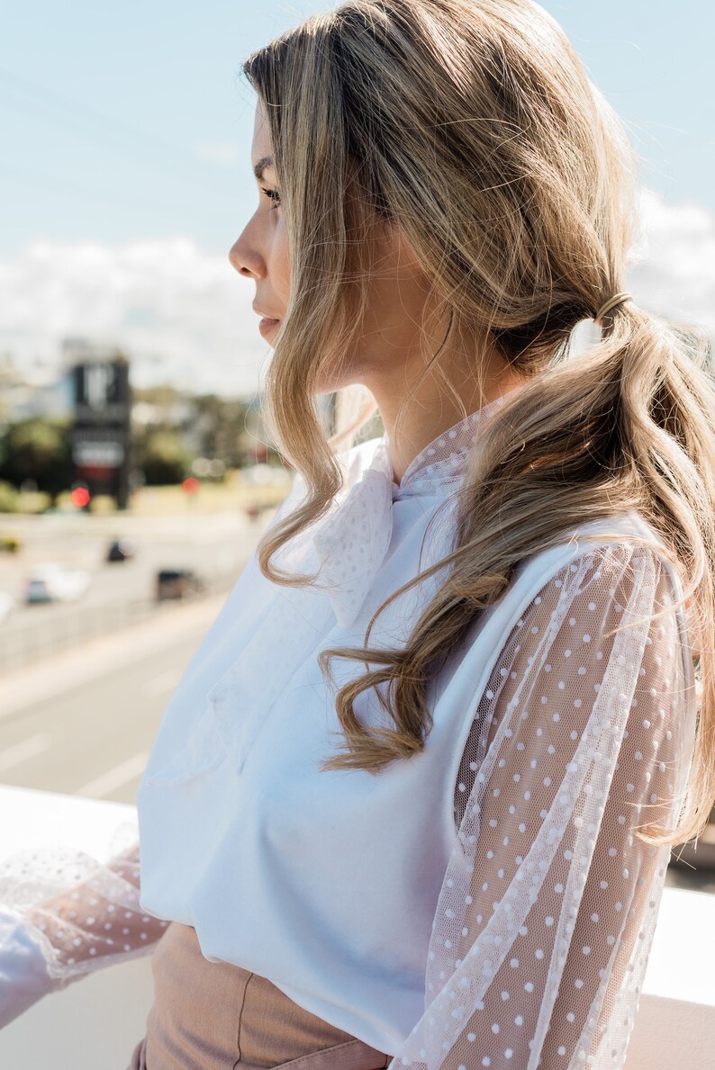 Puede incluir: Una mujer lleva una blusa blanca con una superposici&oacute;n de malla de lunares blancos en las mangas. La blusa tiene un lazo en el cuello. Lleva una falda marr&oacute;n claro.